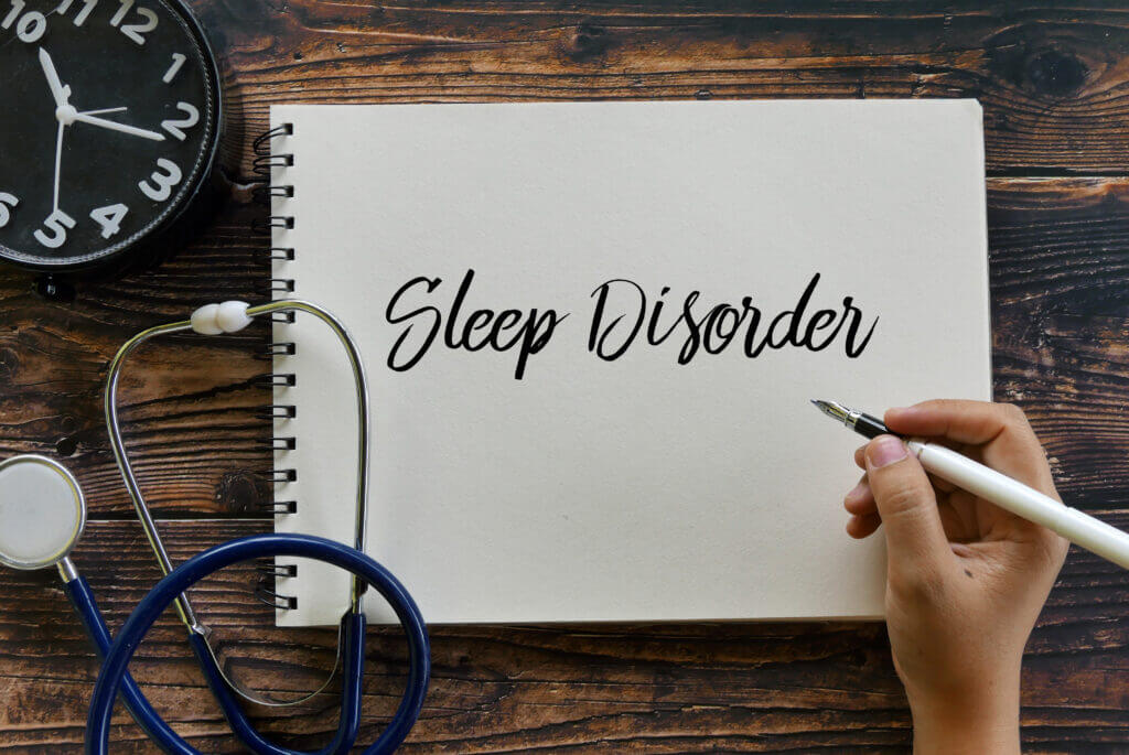Top view of clock, stethoscope, and hand holding pen writing Sleep Disorder on notebook on wooden background.