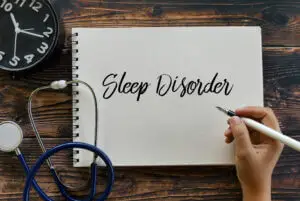 Top view of clock, stethoscope, and hand holding pen writing Sleep Disorder on notebook on wooden background.