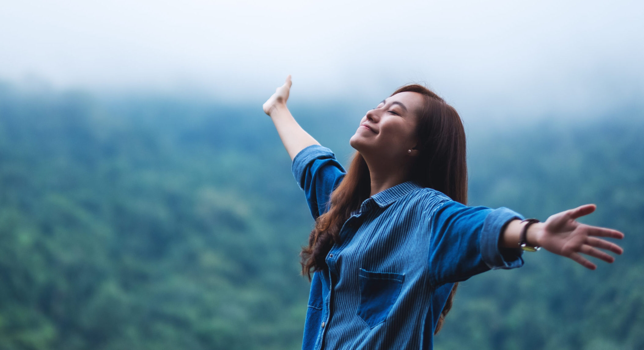 A female traveler close her eyes and open arms with a beautiful green mountains on foggy day in background